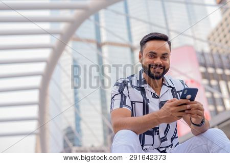 Happy Young Bearded Indian Man With Phone Sitting In The City Outdoors