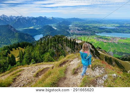 Cute Little Hiker Enjoying Picturesque Views From The Tegelberg Mountain, A Part Of Ammergau Alps, L