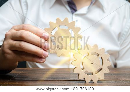 Businessman In White Shirt Connects Two Wooden Gears. Symbolism Of Establishing Business Processes A