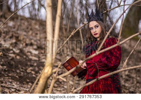 Art Photography. Mysterious Fairy Lady In Red Dress And Black Crown With Old Book. Posing In Forest 