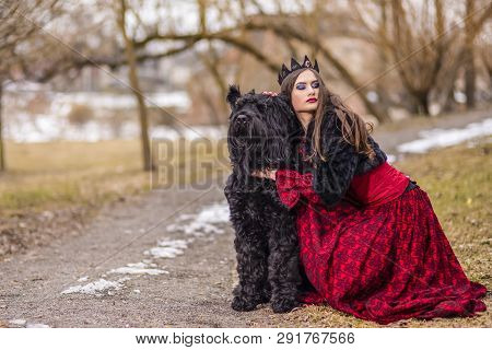Young Princess In Red Dress And Black Fur Jacket Posing In Crown Along With Her Dog In Forest During