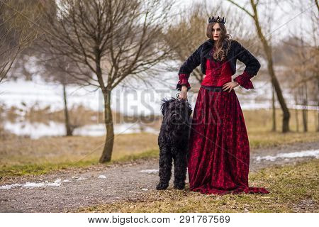 Beautiful Princess In Red Dress And Black Fur Jacket Posing In Crown Along With Her Dog In Forest Du