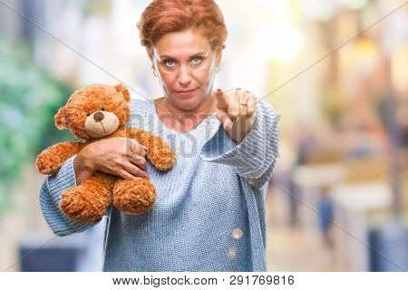 Senior caucasian woman holding teddy bear over isolated background pointing with finger to the camera and to you, hand sign, positive and confident gesture from the front