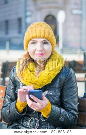 Portrait Of A Young Attractive Woman In A Knitted Hat And Knitted Scarf, Which Sits On A Bench In Th