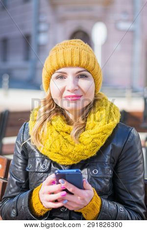 Portrait Of A Young Attractive Woman In A Knitted Hat And Knitted Scarf, Which Sits On A Bench In Th