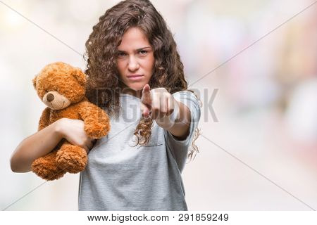 Young brunette girl holding teddy bear over isolated background pointing with finger to the camera and to you, hand sign, positive and confident gesture from the front