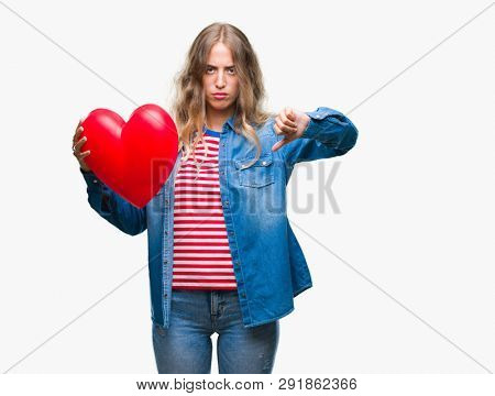 Beautiful young blonde woman holding heart valentine over isolated background with angry face, negative sign showing dislike with thumbs down, rejection concept