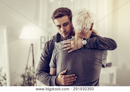 Twenty-years-old Dark-haired Kind Male Calmingly Cuddling Sick Sorrowful Aging Granddad