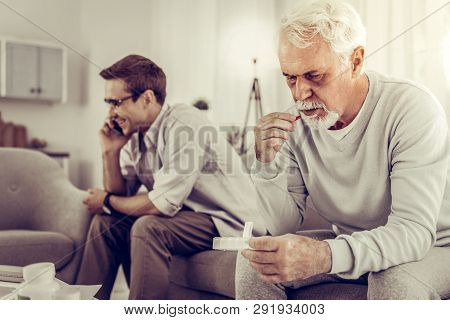 Old Man Taking Pills While Son Indifferently Having A Phone Call.