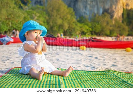 Teething In 8 Month. Baby Girl Sitting On The Beach And Chews A Teether. In Blue Hat And White Dress