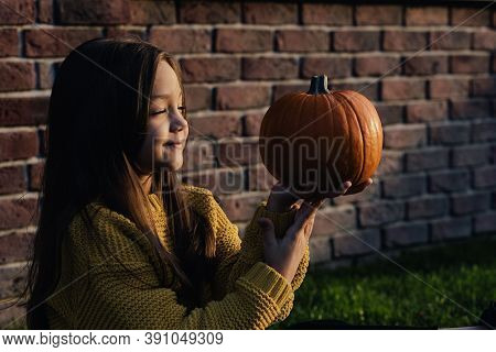 Funny Child Girl In Orange Pullover For Halloween With Pumpkin And On A Dark Brick Background.