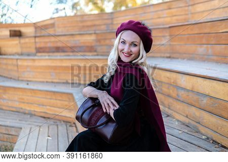 A Smiling Woman Sits On A Summer Theater Bench, Made Of Wood In A Burgundy Coat And Biret, An Adult 
