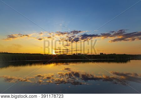 Scenic Summer Sunset Over The Calm Lake Surrounded By Reeds. Golden Clouds Are Reflected In The Wate