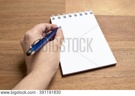 Left Hand Of A Young Left-handed Caucasian Woman Writing With A Blue Pen On A Blank Page Of An Open 