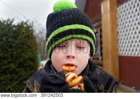 A European Boy In A Warm Jacket And Hat Stands In Winter Near A Fir Tree. The Child Holds The Steeri
