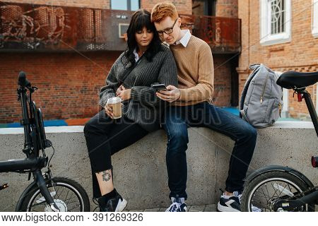 Resting Young Couple, Two Peaople Sitting On A Concrete Border, Looking At Phone