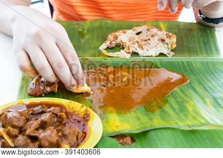 Person Enjoying Eating Roti Canai Served With Curry On Banana Leaf Using Hand