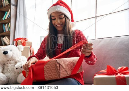 Happy Indian Kid Girl Unties Red Ribbon Opens Christmas Gift Box At Home. Smiling Cute Latin Child W