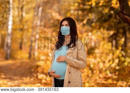 Young Pregnant Woman In Medical Mask On Walk At Autumn Park During Coronavirus Epidemic. Expectant L
