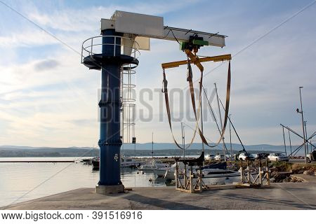 Marine Deck Boat Lifting Crane Mounted On Edge Of Concrete Pier At Local Harbor Surrounded With Calm