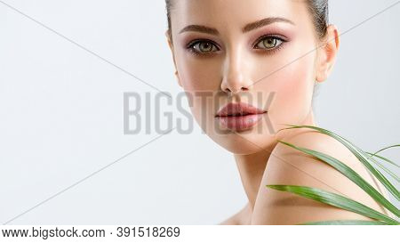 Young beautiful woman with healthy skin of face and palm leaves. Closeup fresh face of an attractive caucasian girl with green plants. Model with bright brown eye makeup. Skin care concept. 