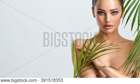 Young beautiful woman with healthy skin of body and palm leaves. Tanned body of an attractive white girl with green plants. Woman hiding her naked body with hands. Large palm leaves cover the body.