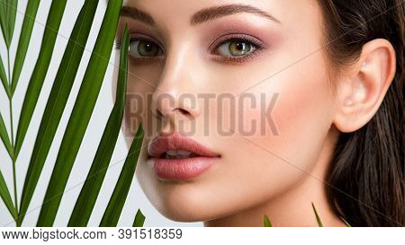 Young beautiful woman with healthy skin of face and palm leaves. Closeup fresh face of an attractive caucasian girl with green plants. Model with bright brown eye makeup. Skin care concept. 