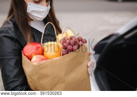 Close Up Of Groceries In Females Hand By The Car Truck. Woman In Protective Mask And Disposable Glov