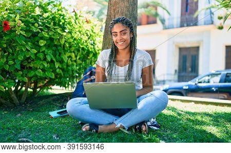 Young african american student woman smiling happy using computer laptop sitting on the grass at the university campus
