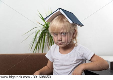 Fair-haired Boy Sits With Book On His Head. Angry Little Boy Holding Book Above His Head Like Roof.