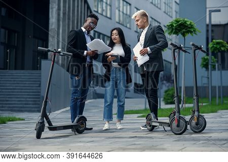Expirienced International Business Team Of Three Co Workers Analyzes Project Standing Near E-scooter