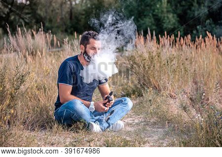 Brutal Vaper With Electronic Smoke Device On The Grass Of The Forest. The Man Smokes.