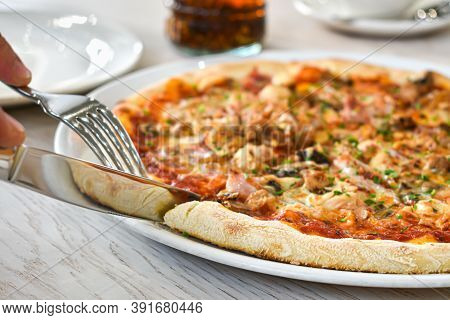 Hands With A Knife And Fork Cut A Slice Of Pizza. Fresh Pizza On A White Dish, Restaurant Interior. 