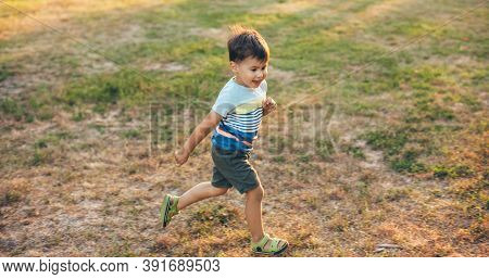 Running Caucasian Boy Is Smiling On A Green Field In A Park During A Summer Walk