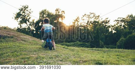 Small Boy On Bike Is Riding And Cheering In A Park On A Green Field During A Sunset