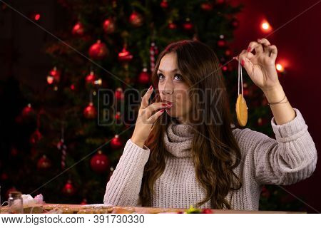 Portrait Of Pretty Brunette Girl Licks Her Fingers Decorating Christmas Cookies On Dark Blurred Back