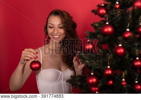 Smiling Long-hair Ginger Girl In Open Shoulders Dress Looking At Toy Ball On Christmas Tree Dressing