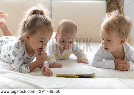 Three Little Smiling Kids Play Together On Soft White Bed At Home. Brother And Siste Read Book Fairy
