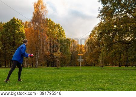 Young Caucasian Man In Blue Jacket Playing Disc Golf On Autumn Play Course With Basket