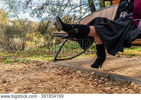 A Cheerful Lady Sits On A Garden Swing, Forged In A Burgundy Coat And Biret, An Adult, In The Fall A