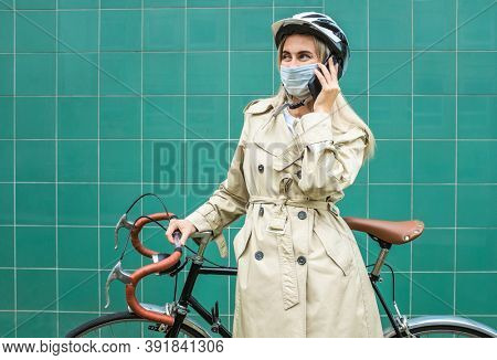 Woman in protective maskin, in helmet using her smartphone device. Female go to work on his sport retro bike. Girl standing on green background.