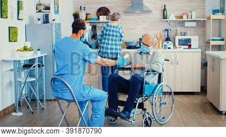 Geriatrician Giving Pills To Elderly Disabled Patient In Wheelchair During Coronavirus Home Visit. M
