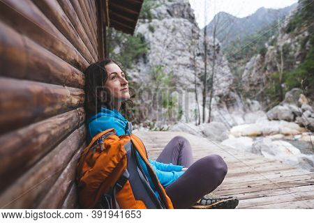 A Woman With A Backpack Sits On The Porch Of An Old Wooden House. The Traveler Found A Hut For An Ov