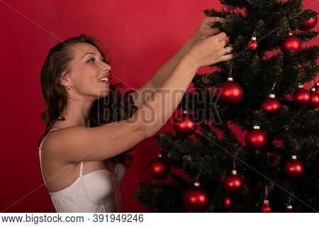 Happy Young Pretty Woman In Festive Dress Decorating Xmas Tree On Red Background. Christmas Music So
