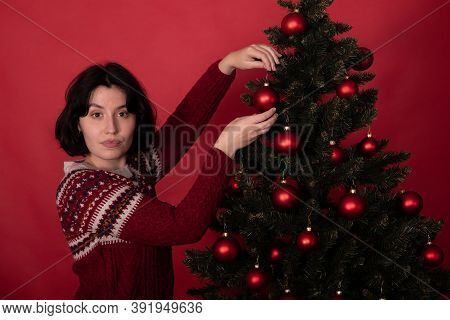 Young Pretty Woman In Knitted Traditional Sweater With Ornament Decorating Xmas Tree Looks At Camera