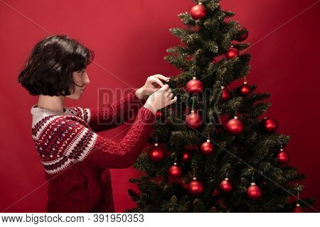 Young Pretty Woman In Knitted Traditional Sweater With Ornament Decorating Xmas Fir Tree On Red Back