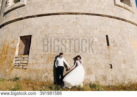 Bride And Groom Are Holding Hands And Standing Tjgether Near The Wall Of The Arza Fortress
