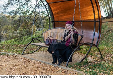 Sits On A Garden Swing, Forged In A Burgundy Coat And Biret, An Adult With A Beautiful Smile, In The