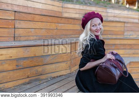 Cheerful Sits On A Summer Theater Bench, Wooden In A Burgundy Coat And Biret, An Adult Smiles At The