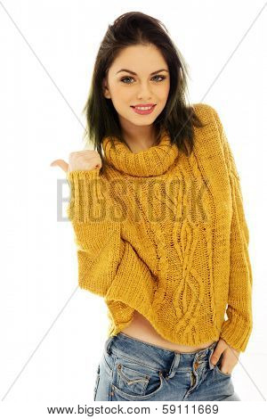 Trendy beautiful young woman in a gold pull-over and jeans giving a thumbs up of approval as she smiles at the camera, on white
